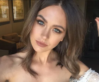 A woman with brown hair and subtle makeup, wearing an off-shoulder top, looks into the camera in a softly lit room.