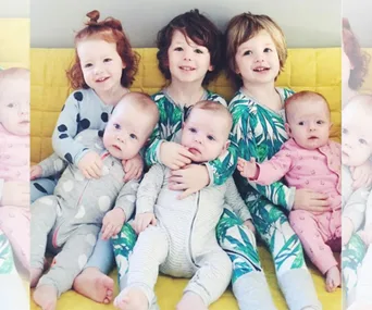Young children and babies sit together on a yellow couch, wearing colorful pajamas.