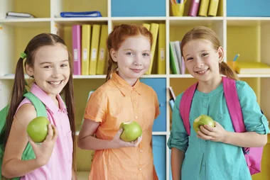 Three smiling schoolgirls holding apples, standing in front of colorful shelves.