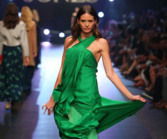 Model in a flowing green dress walks down a dimly lit runway, with audience and other models in the background.