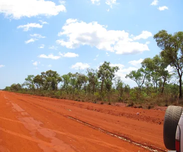 Red dirt road with green trees and blue sky in rural Australia. A car tire is visible on the right.