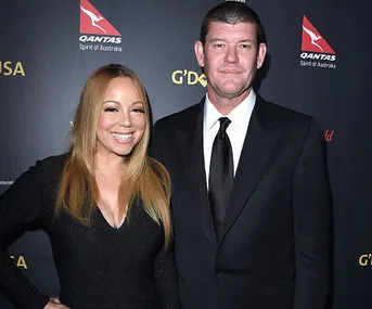 Mariah Carey and James Packer smiling together at a formal event, both in black attire, against a branded backdrop.