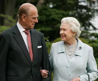 A distinguished elderly couple smiles lovingly at each other, walking arm-in-arm in an outdoor setting.