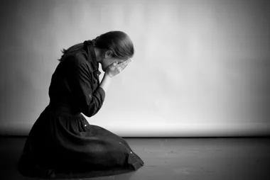 A woman kneeling, covering her face with her hands, in a monochrome setting, conveying sadness or distress.
