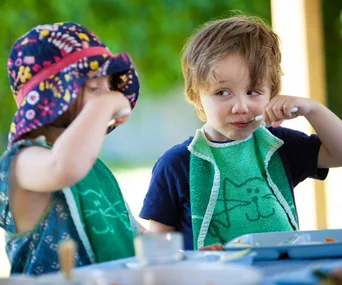 Children eating outdoors, wearing colorful bibs; one with a floral hat, both using spoons.