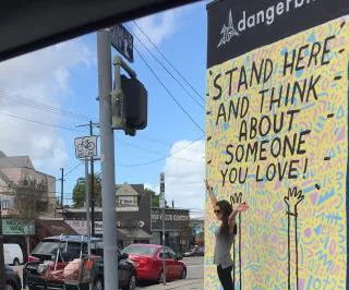 Person poses playfully against a colorful mural that reads "Stand here and think about someone you love!" in an urban area.