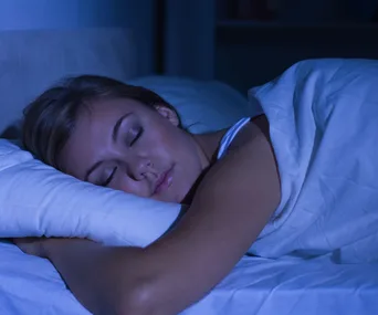 A woman sleeping peacefully on her side in a dimly lit room, covered with a white blanket.