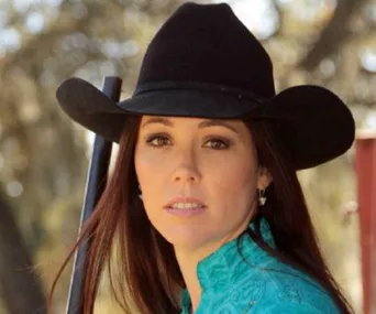 Person wearing a black cowboy hat and turquoise shirt outdoors, holding a rifle over their shoulder.