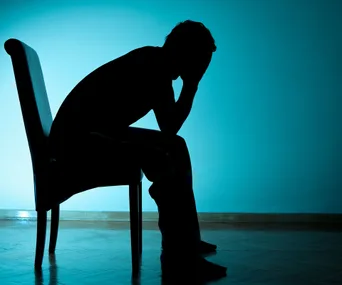 A silhouette of a person sitting on a chair, head in hands, against a blue background.