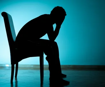 A silhouette of a person sitting on a chair, head in hands, against a blue background.