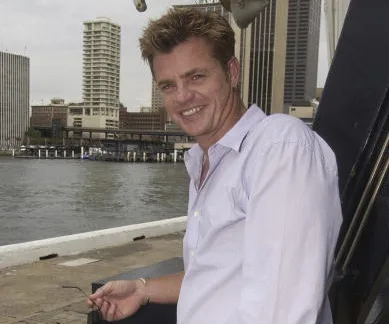 A man in a light shirt leans against a railing by the waterfront, with city buildings in the background.