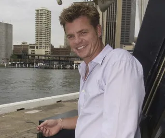 A man in a light shirt leans against a railing by the waterfront, with city buildings in the background.