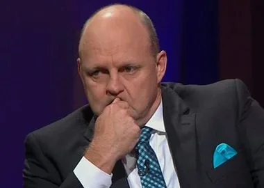 Man in a suit looks thoughtful with hand on chin, blue tie and pocket square, against a dark background.