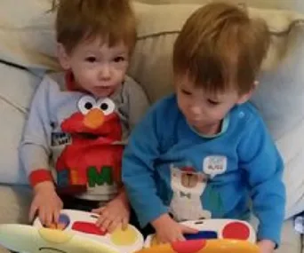 Two toddlers sitting on a couch playing with a colorful toy, wearing shirts with playful designs.