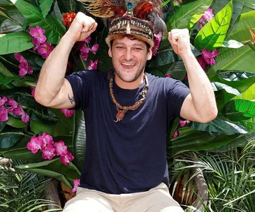Person celebrating with hands raised, wearing feathered headpiece, surrounded by tropical plants and pink flowers.