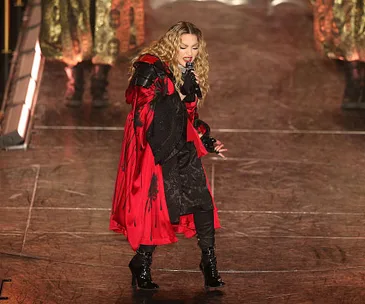 Singer performing energetically on stage in a red and black outfit, surrounded by dancers.