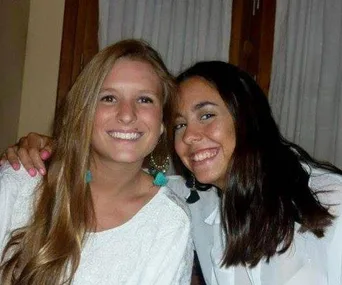 Two smiling young women, one with long blonde hair and the other with dark hair, pose closely together indoors.