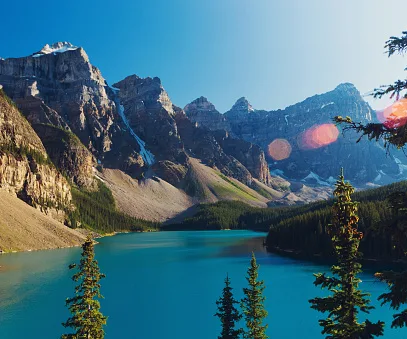 A serene mountain landscape with a blue lake surrounded by lush evergreens and rocky peaks under a clear sky.