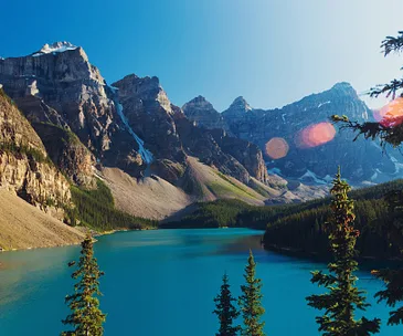 A serene mountain landscape with a blue lake surrounded by lush evergreens and rocky peaks under a clear sky.
