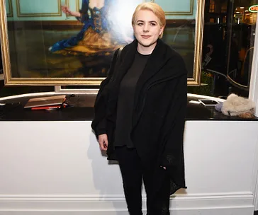 Person in black outfit standing indoors in front of a large framed painting.