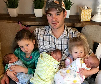 Father sitting with two young daughters and three swaddled newborn triplets on a couch.