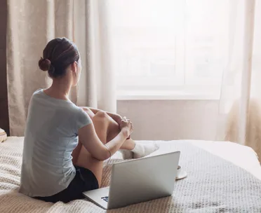 A woman sits on a bed near a laptop, gazing out a window with earbuds in, suggesting introspection or relaxation.
