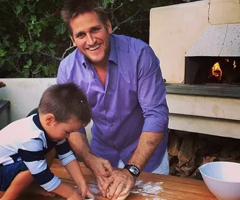Man and young child smiling while kneading dough outdoors near a stone oven.
