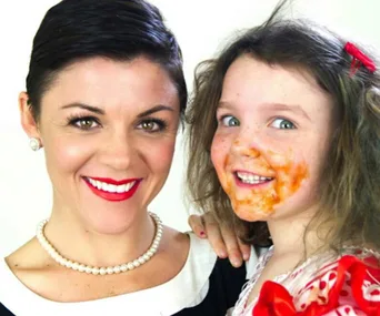 Women in pearls and red-lipped smile with a young child in a stained outfit and messy hair, both looking at the camera.