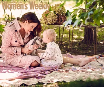 Kate Ritchie sits with her baby, Mae, on a blanket in a garden, holding a small rabbit under a tree.