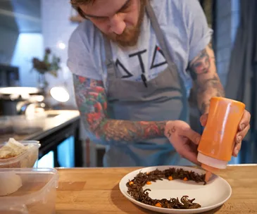 Chef applying sauce onto a plate of edible insects in a kitchen setting.