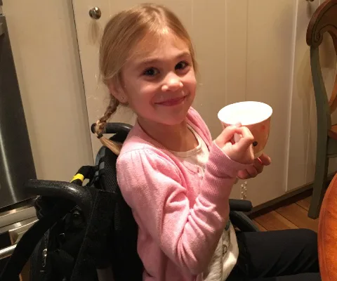 Young girl in a wheelchair, smiling and holding a cup, with a pink cardigan in a kitchen setting.