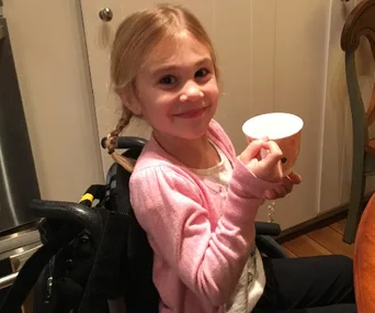 Young girl in a wheelchair, smiling and holding a cup, with a pink cardigan in a kitchen setting.