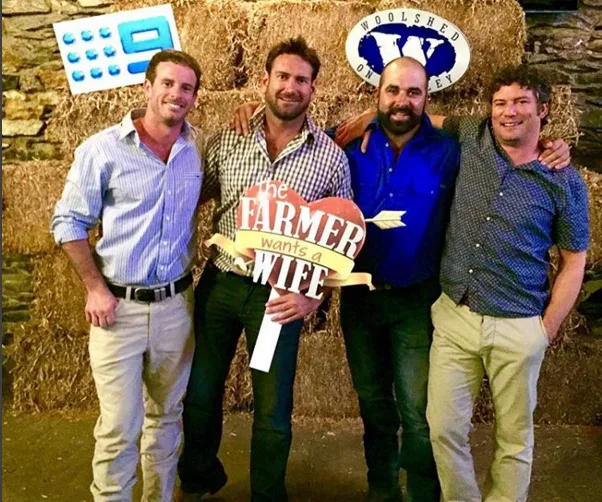 Four men posing in front of hay bales, one holding a "The Farmer Wants a Wife" sign.