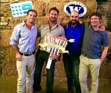 Four men posing in front of hay bales, one holding a "The Farmer Wants a Wife" sign.