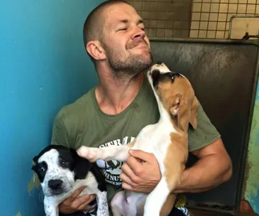 Man smiling with two puppies, one licking his face affectionately.