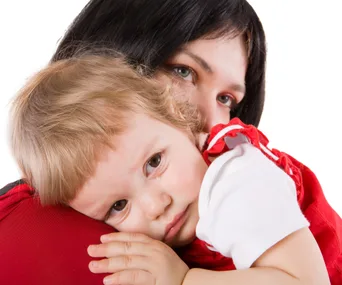 Child resting head on woman's shoulder, appearing sad, wearing a red and white outfit.