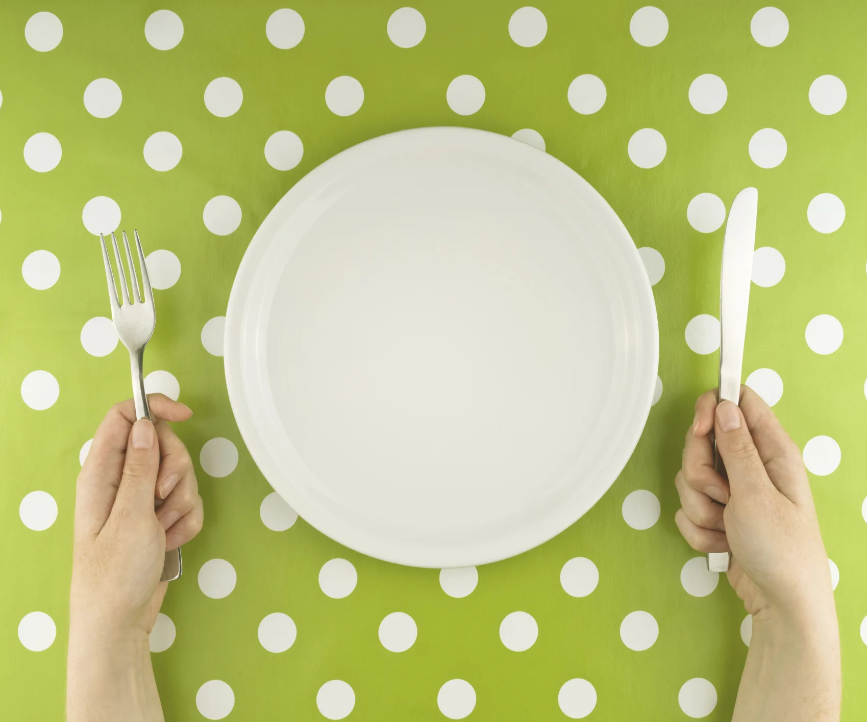Empty plate on green polka dot tablecloth, hands holding fork and knife.