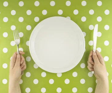 Empty plate on green polka dot tablecloth, hands holding fork and knife.