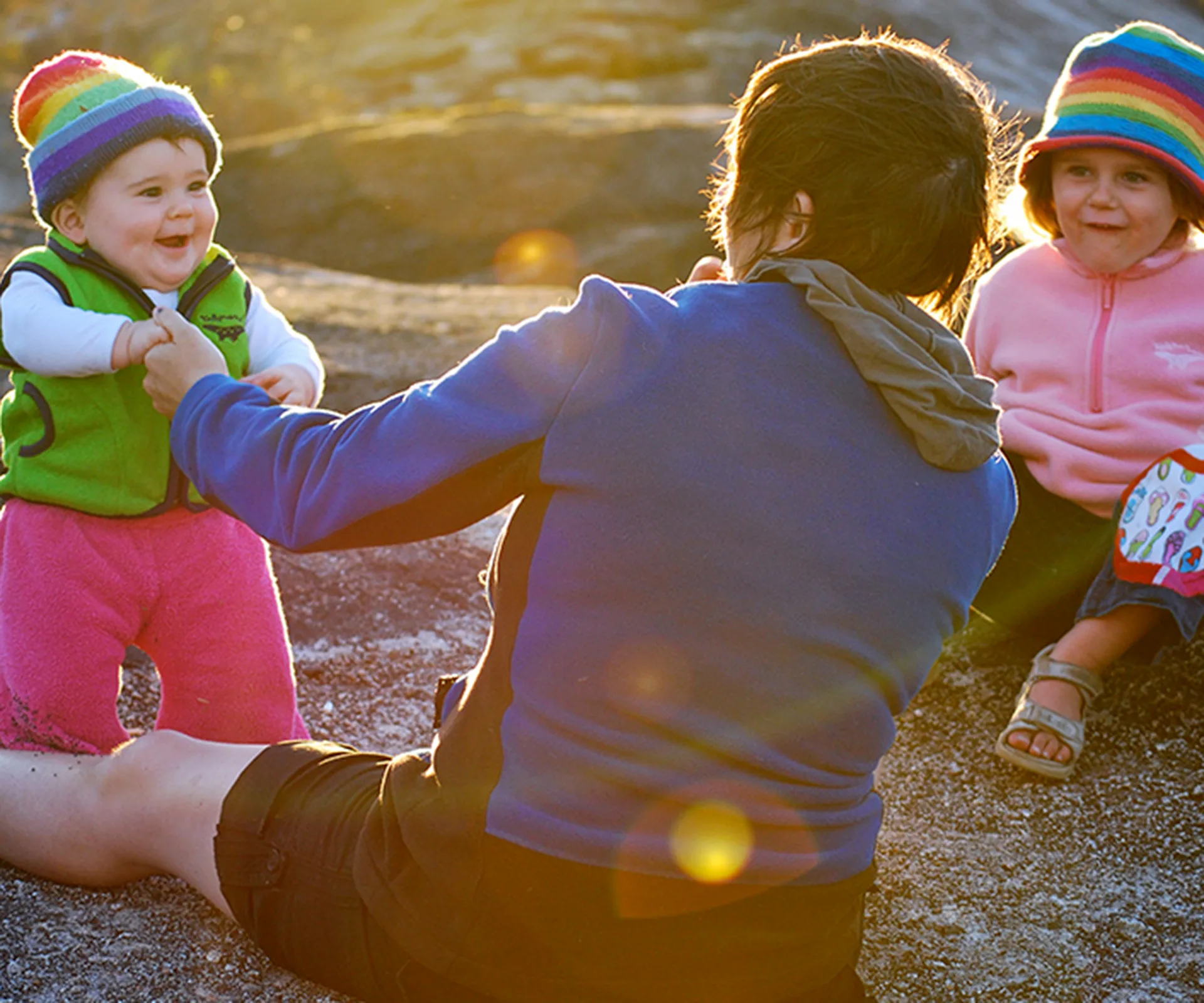 A parent interacts with two kids in colorful hats outdoors, one standing and one sitting on the ground.