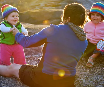 A parent interacts with two kids in colorful hats outdoors, one standing and one sitting on the ground.