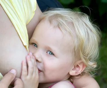Toddler with blond hair and blue eyes breastfeeding, partially visible adult in yellow clothing, close-up.