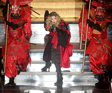 Madonna performs on stage wearing a red and black costume, flanked by dancers in red outfits, holding poles.