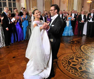 A couple elegantly dances in a formal ballroom, surrounded by an applauding crowd in evening attire.