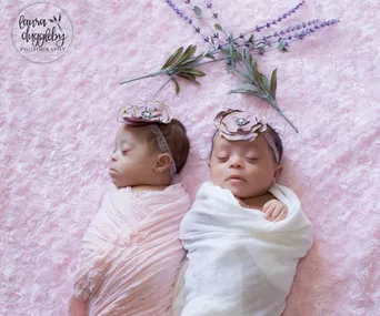 Twin babies with Down syndrome sleeping, wrapped in pastel blankets with floral headbands on a pink textured blanket.