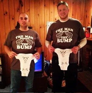 Two men wearing "The Man Behind The Bump" shirts holding baby onesies at a dad-chelor party in a wooden room.
