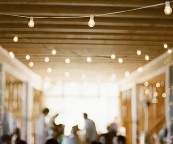 Blurred indoor wedding scene with string lights and indistinct figures in the background.