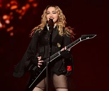 Madonna performing on stage with a guitar, wearing a black outfit and smiling while singing into a microphone.