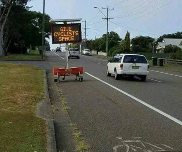 "Electronic road sign reads 'GIVE CYCLISTS SPACE' beside a road with a bicycle lane and passing vehicles."