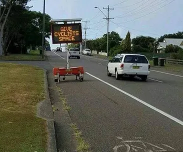 "Electronic road sign reads 'GIVE CYCLISTS SPACE' beside a road with a bicycle lane and passing vehicles."
