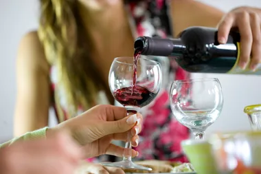 A person pours red wine into a glass held by another person, with an empty glass nearby.